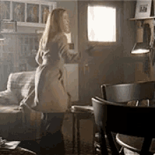a woman in a trench coat is standing in a living room next to a chair and a table .