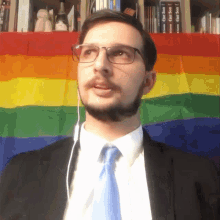 a man wearing glasses and a tie stands in front of a rainbow flag with books on the shelf behind him