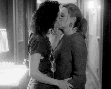 two women are kissing in a hallway with a lamp in the background