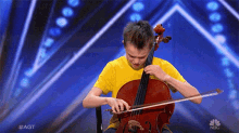 a young man in a yellow shirt is playing a cello on a stage with a nbc logo in the background