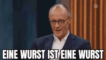 a man in a suit and glasses is saying eine wurst ist eine wurst while sitting on a stage .