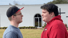 a man wearing a hat with a cross on it stands next to another man in a red hoodie