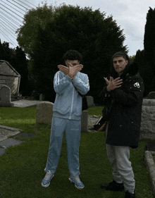 two young men standing in a cemetery with one wearing a light blue tracksuit