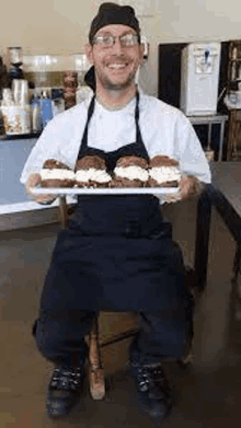 a man in an apron is sitting on a chair holding a tray of desserts .