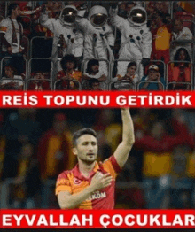 a soccer player in a red and orange jersey stands in front of a crowd with a sign that says reis topunu getirdik