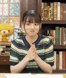 a girl is sitting at a table with her hands folded in front of a bookshelf with a teddy bear in the background .