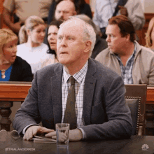 a man in a suit and tie is sitting at a table with a glass of water in front of a crowd of people .