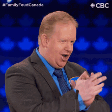 a man in a suit and tie applauds in front of a microphone with the words familyfeud canada behind him