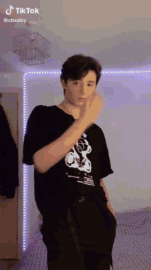 a young man in a black t-shirt and black pants is standing in a room with his hand in his pocket .