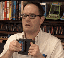 a man wearing glasses is holding a mug in front of a shelf of video games including sonic the hedgehog