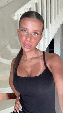 a woman in a black tank top is taking a selfie in front of a set of stairs .