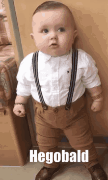 a baby wearing suspenders and a white shirt has the name hegobald written on the bottom