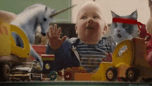 a baby is sitting on a table surrounded by toys and a cat wearing a bandana .