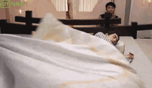 a man is laying on a bed with a white blanket while another man looks on .