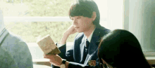 a boy in a suit and tie is reading a book in a classroom while sitting next to a girl .