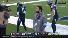 a man is standing on the sidelines of a football game while two football players are running on the field .