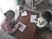 two girls sit at a table with their faces blurred and one of them is writing