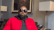 a man wearing sunglasses and a red jacket is sitting in a living room .