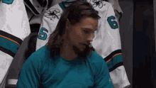 a man with long hair and a beard is sitting in a locker room in front of a hockey jersey .