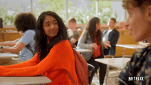 a girl in an orange sweater sits at a desk in a classroom with a netflix logo on the bottom