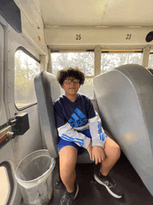 a boy wearing a blue adidas shirt sits in the back seat of a bus