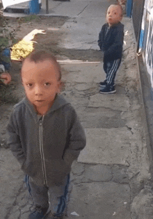 two little boys are standing next to each other on a sidewalk and one of them is looking at the camera .