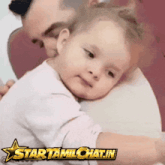 a man is holding a baby in his arms with a star tamilchat.in logo behind him
