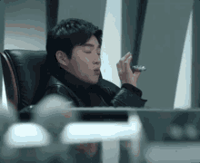 a man in a leather jacket is sitting at a desk holding a cell phone in his hand .