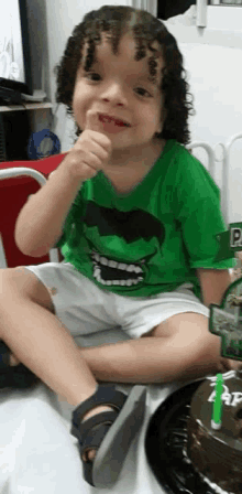 a young boy wearing a green hulk shirt is sitting next to a birthday cake .