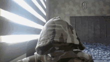 a person wearing a camouflage hoodie is sitting in front of a window with blinds