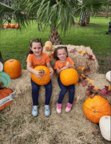 two little girls are sitting on a bale of hay holding pumpkins and wearing orange shirts that say welcome