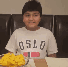 a boy wearing a white oslo shirt sits at a table with a bowl of chips