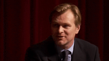 a man in a suit and tie is standing in front of a red curtain and smiling .