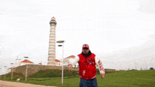 a man in a red and white jacket stands in front of a grassy field with the ocean in the background