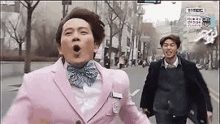 a man in a pink suit and bow tie is running down a street next to another man .