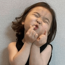 a little girl is making a funny face with her eyes closed and her hands in her mouth .