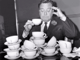 a man in a suit and tie is sitting at a table with stacks of cups and saucers