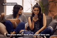 two women are sitting on a couch looking at a cell phone and talking to each other .