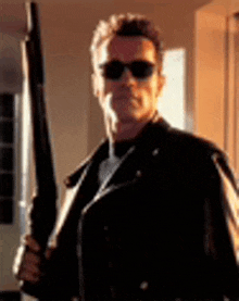 a man wearing sunglasses and a leather jacket is holding a gun in a room .