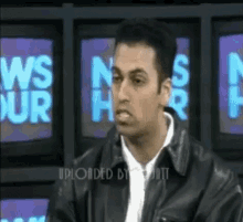 a man in a leather jacket is sitting in front of a wall of television screens with the words news hour written on them