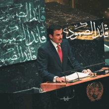 a man in a suit and red tie stands at a podium with arabic writing on the wall behind him