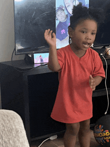 a little girl in a red shirt stands in front of a tv and a playground ball