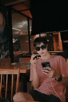 a man wearing sunglasses and a pink shirt is sitting at a table using his phone