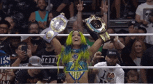 a woman with green hair is holding two championship belts in a wrestling ring in front of a crowd .