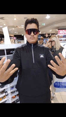 a man wearing sunglasses and an adidas jacket holds his hands out