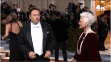 a man in a tuxedo and a woman in a pearl necklace are standing next to each other on a red carpet