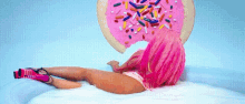 a woman with pink hair is laying on a bed next to a donut with sprinkles .