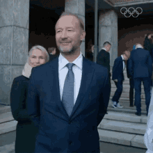 a man in a suit and tie stands in front of a building with the olympic rings on the wall