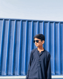 a boy wearing sunglasses and a black shirt stands in front of a blue wall