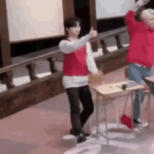 a man in a red vest is giving a thumbs up while standing in front of a desk in a classroom .